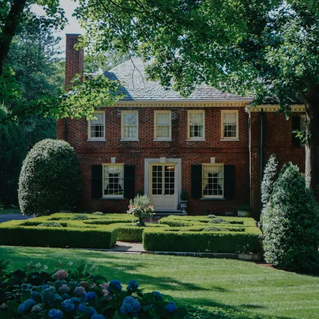 A brick colonial home behind a trimmed hedge garden